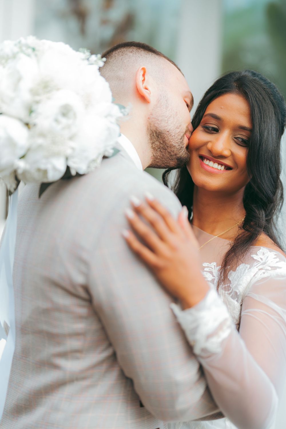 A groom in a gray suit kisses his smiling bride in a white lace dress while holding a white floral bouquet.