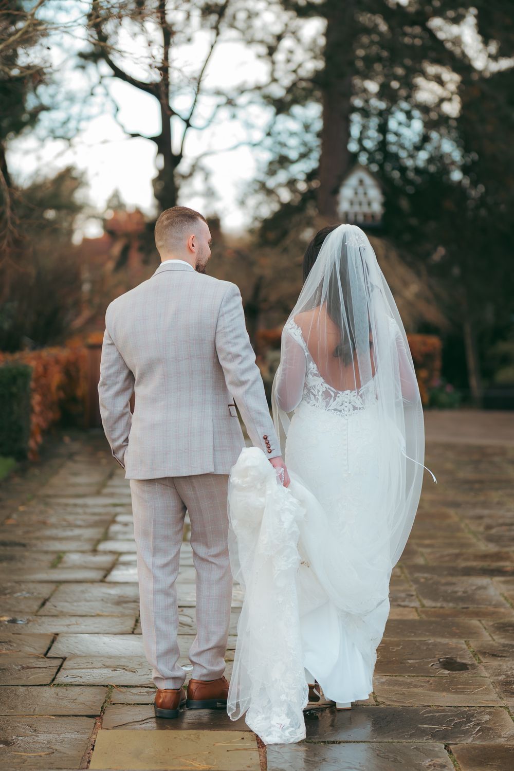 A bride and groom walk hand-in-hand down a stone path, viewed from behind, surrounded by autumn trees.