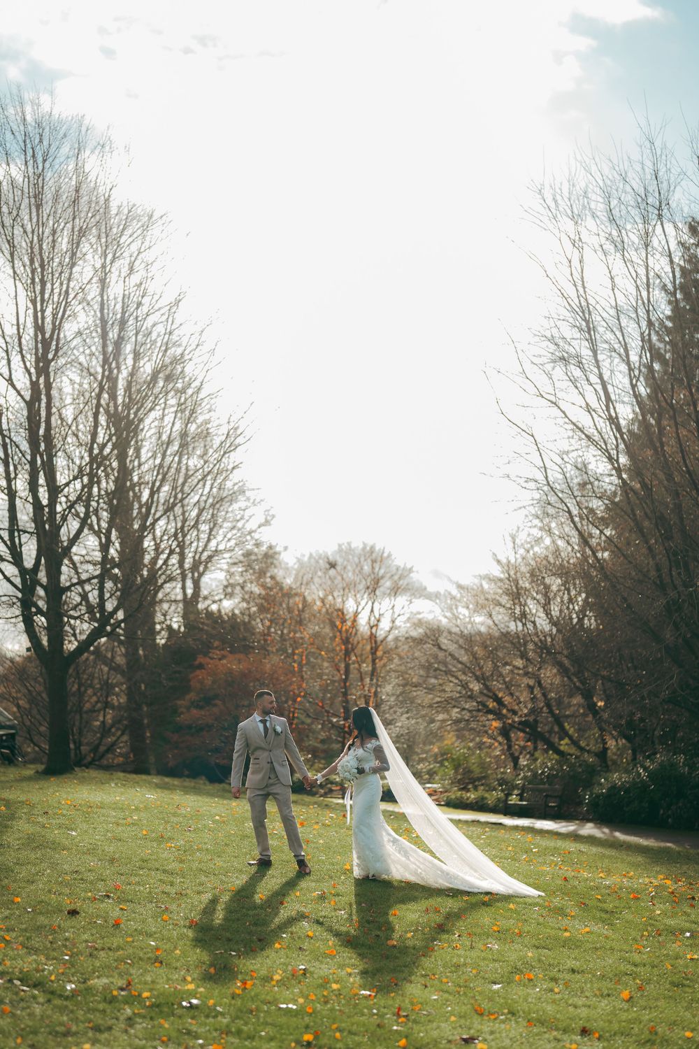 Bride and groom holding hands on a green lawn surrounded by autumn trees on their wedding day.