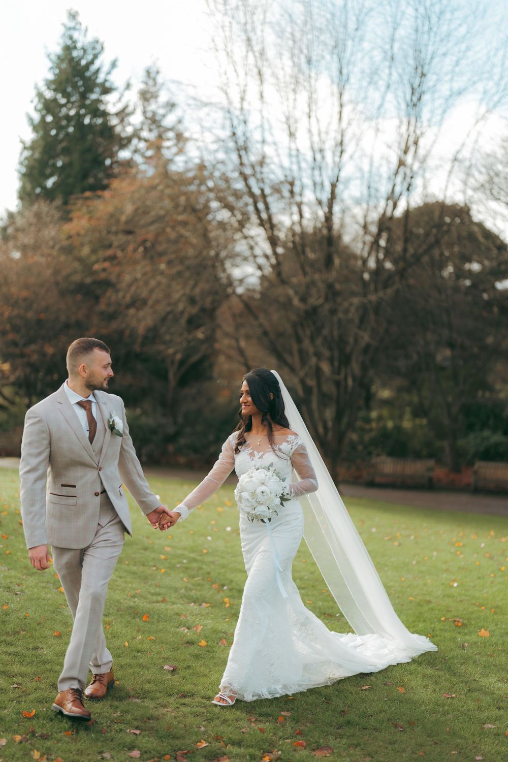 Bride and groom walk hand in hand across lush green lawn, she in lace gown with veil, he in light gray suit.