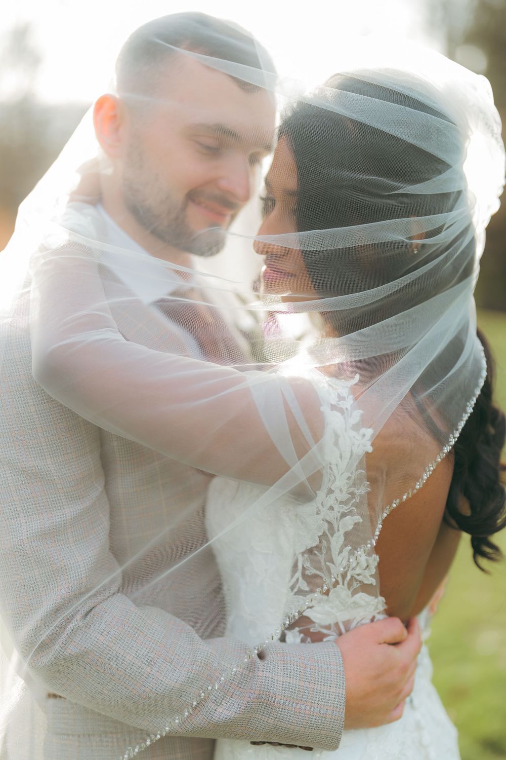 Bride and groom embracing under a sheer veil in golden sunlight, wearing lace wedding dress and gray suit.