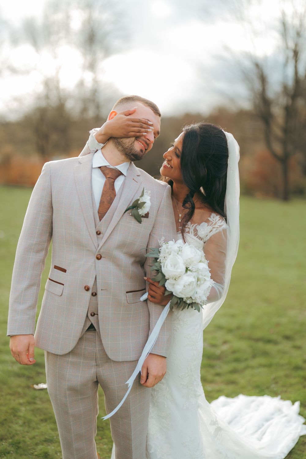 Bride playfully covers groom's eyes while holding white bouquet, both smiling outdoors in autumn setting.