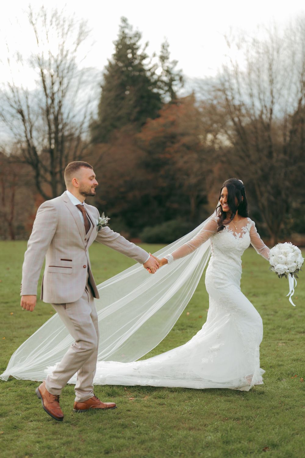 Bride and groom holding hands walking on green grass outdoors, bride in white lace gown with flowing veil, groom in beige suit.