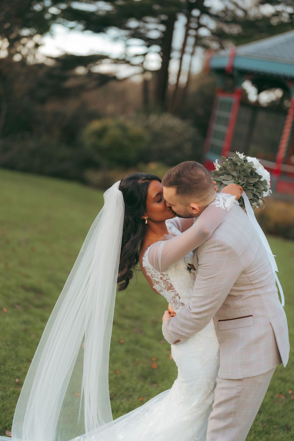 Bride and groom share a romantic kiss outdoors, her white veil flowing as she holds a bouquet in a lush garden setting.