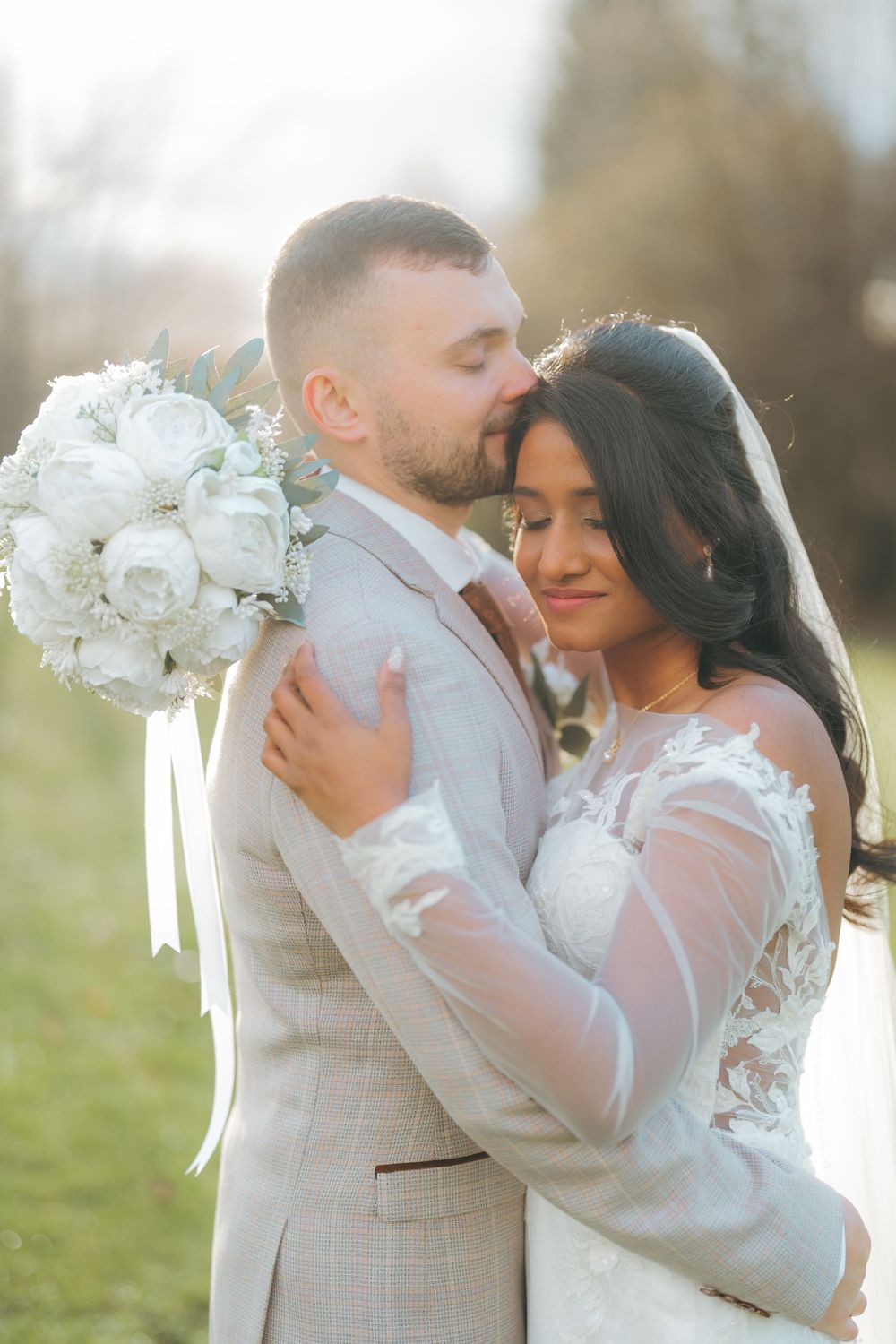 A groom in a light gray suit kisses his bride wearing a white lace gown and veil while holding a white rose bouquet.