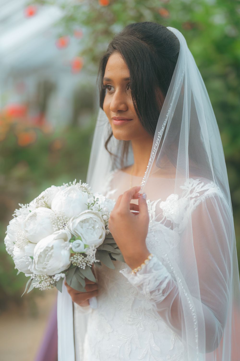 Bride in white lace gown and veil holding white peony bouquet, posing outdoors with soft floral background.