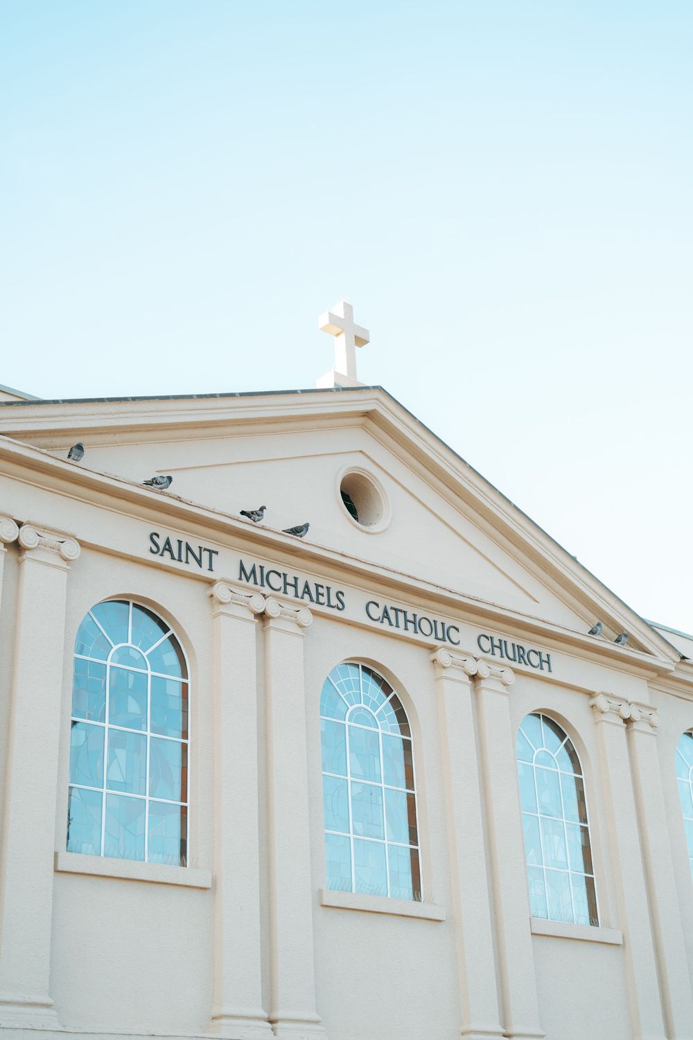 Saint Michaels Catholic Church facade with arched windows, cross atop pediment, and birds perched on the cornice against a clear sky.