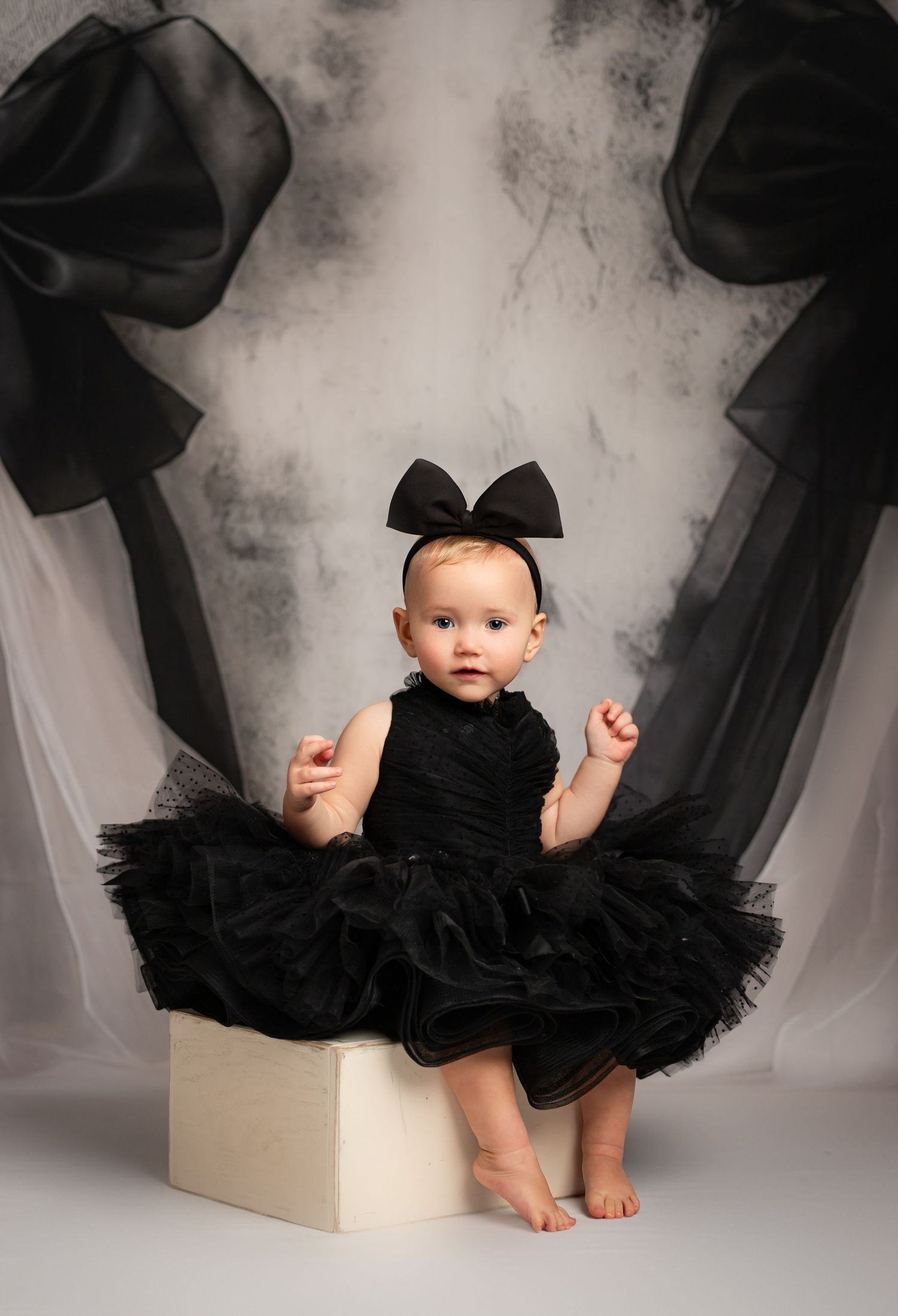 Baby girl in black tutu dress and bow headband sits on white block before elegant gray backdrop with black bows.