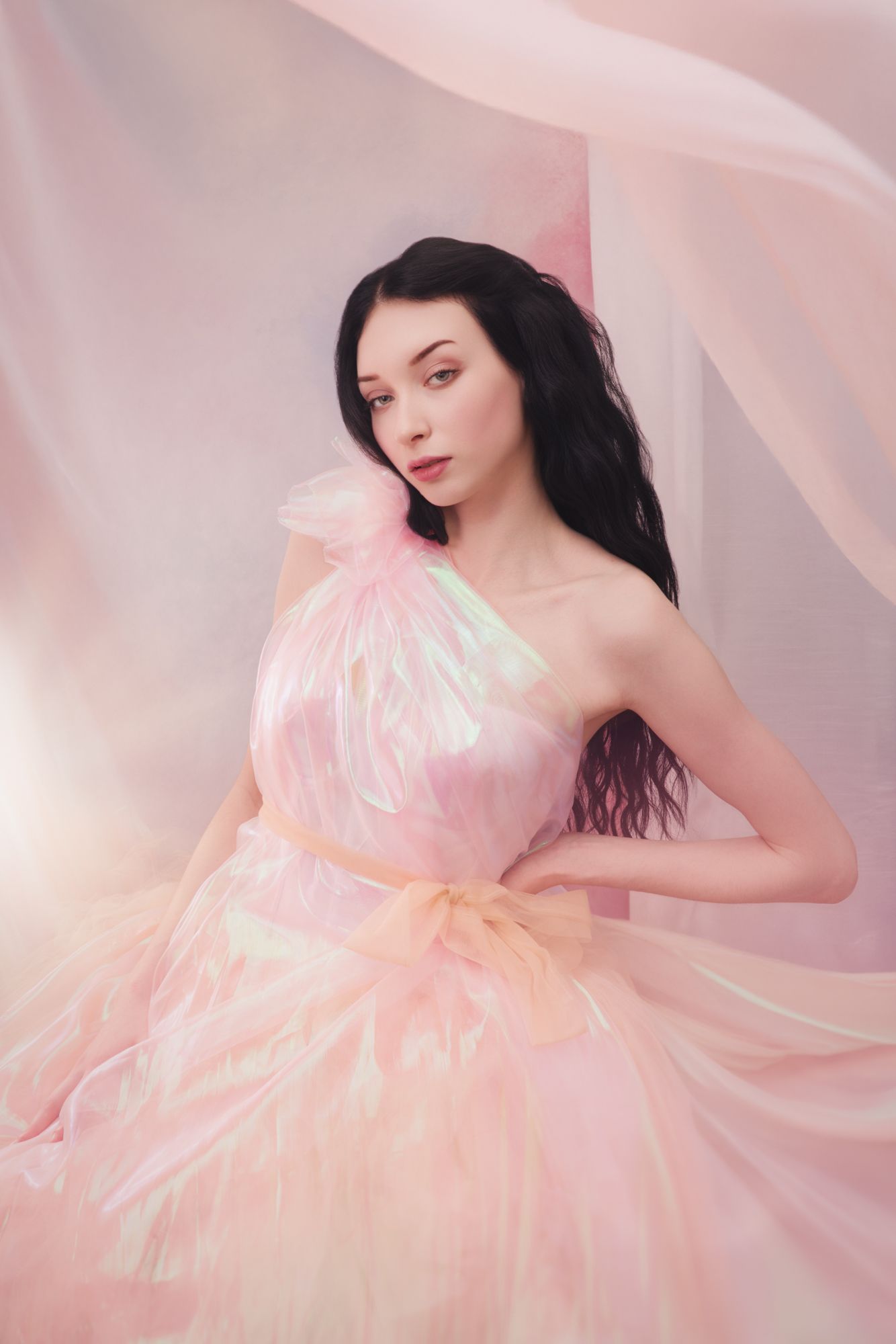 Young woman in a flowing iridescent pink tulle gown poses gracefully against soft pink draped fabric backdrop.