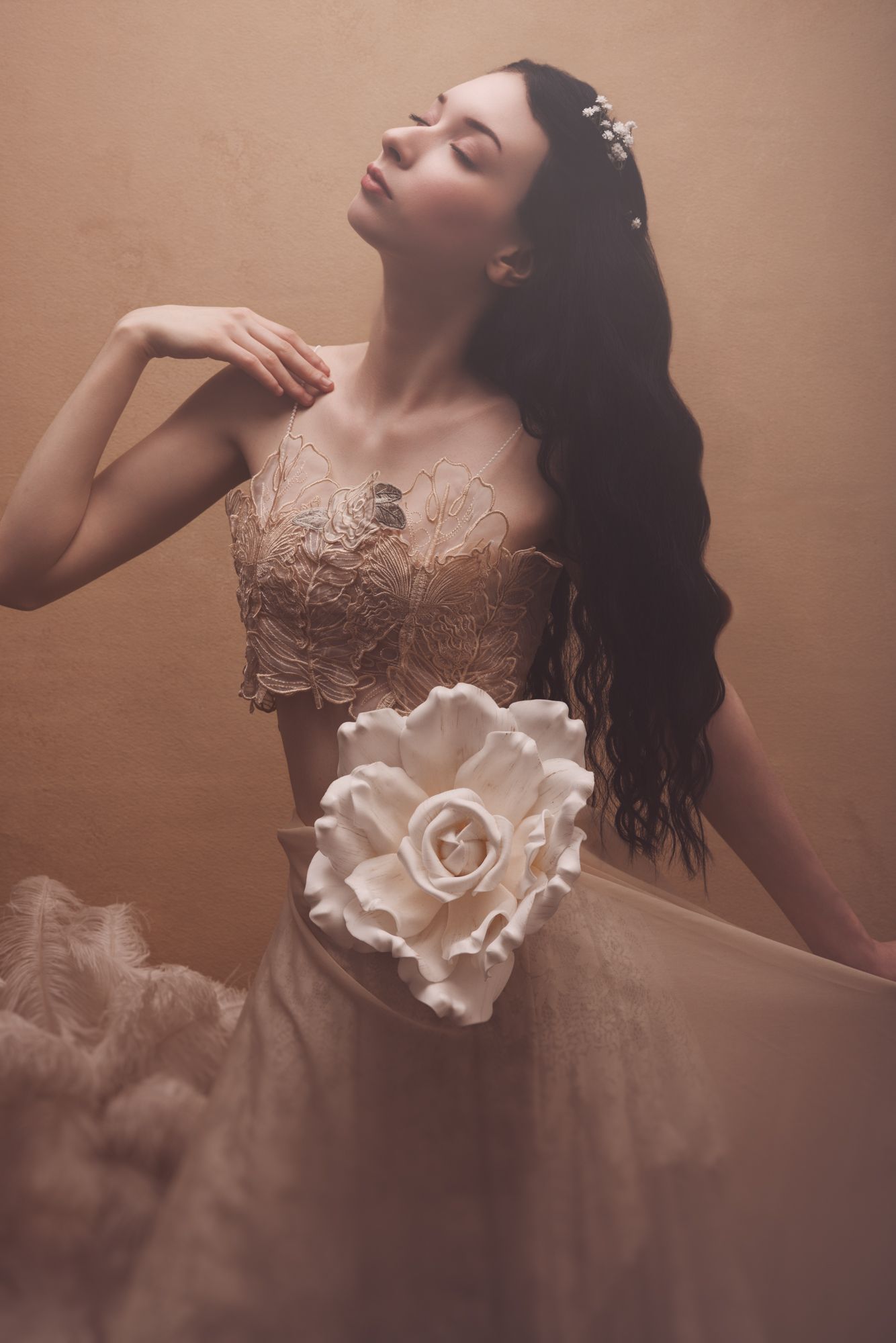 Elegant woman in beige lace corset gown with large rose detail, dark wavy hair, eyes closed, posing gracefully.