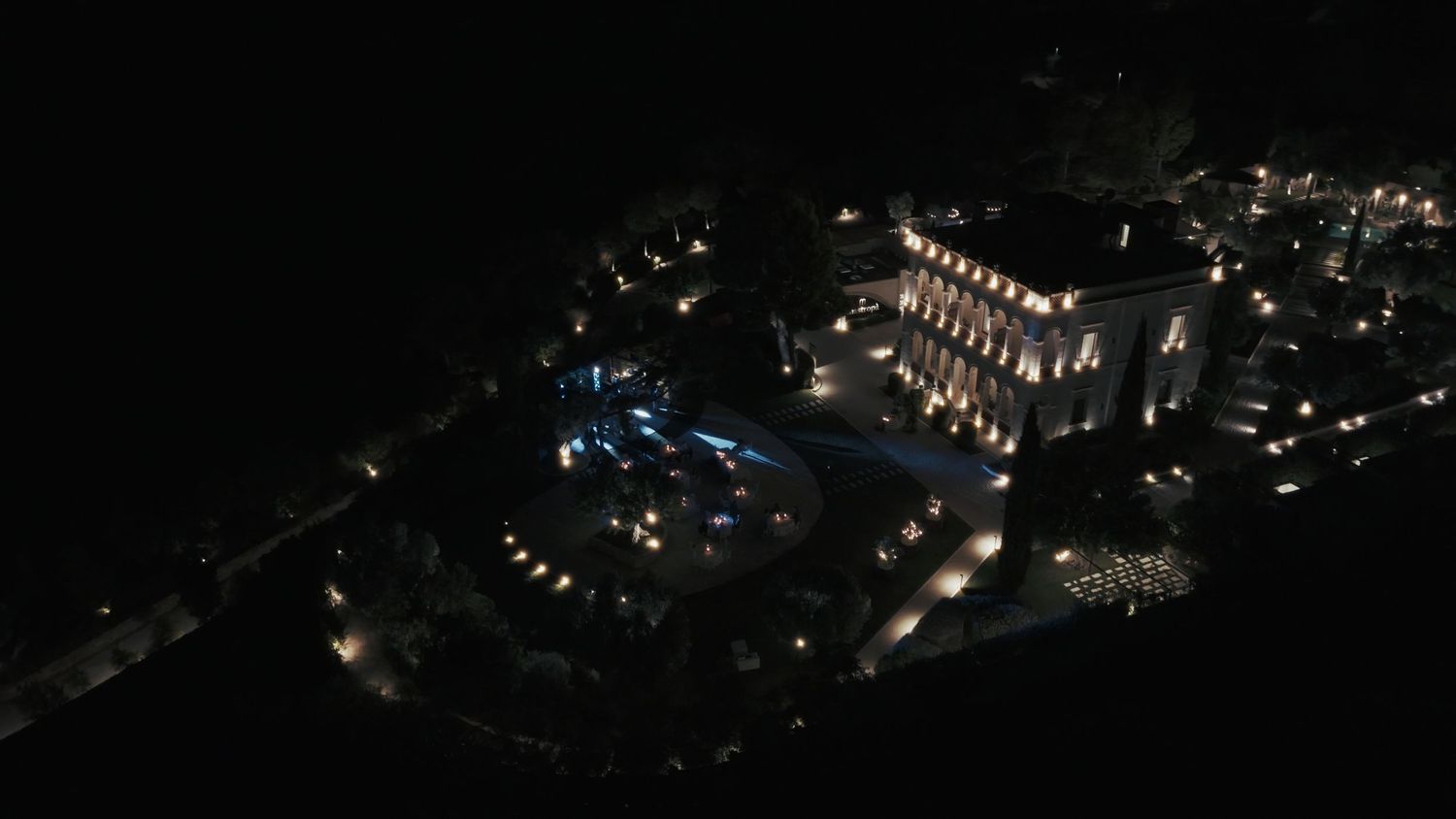 Aerial night view of an illuminated luxury resort with pool and landscaped grounds glowing against the dark sky.