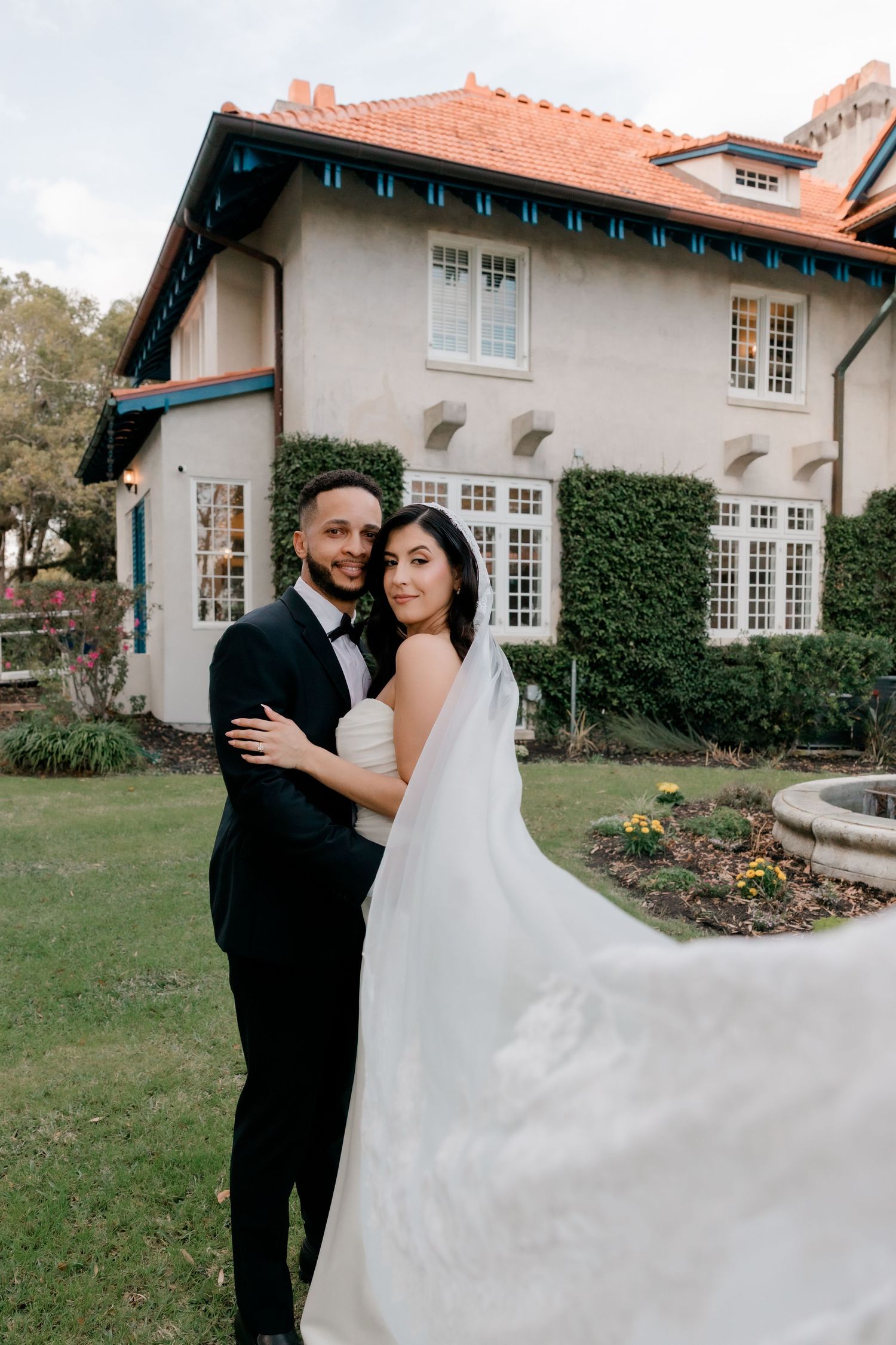 A bride and groom embrace in front of an elegant Spanish-style estate with red tile roof and lush ivy walls.