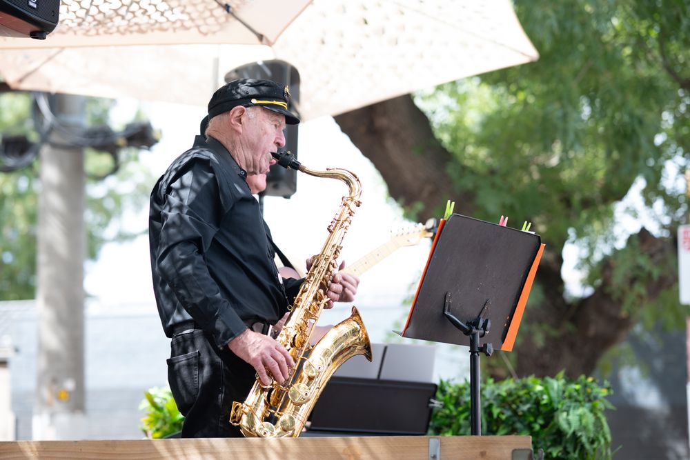 Elderly musician playing saxophone on an outdoor stage under a white umbrella at a live event.
