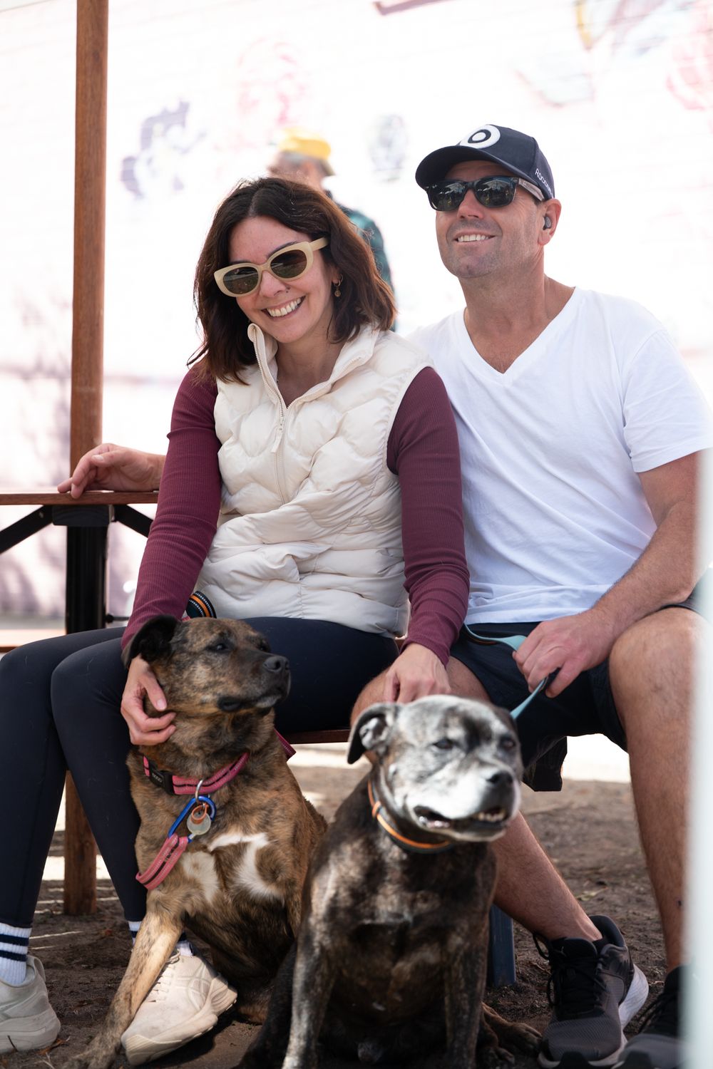 Smiling couple sitting outdoors with two brindle dogs on leashes at a casual pet-friendly venue.