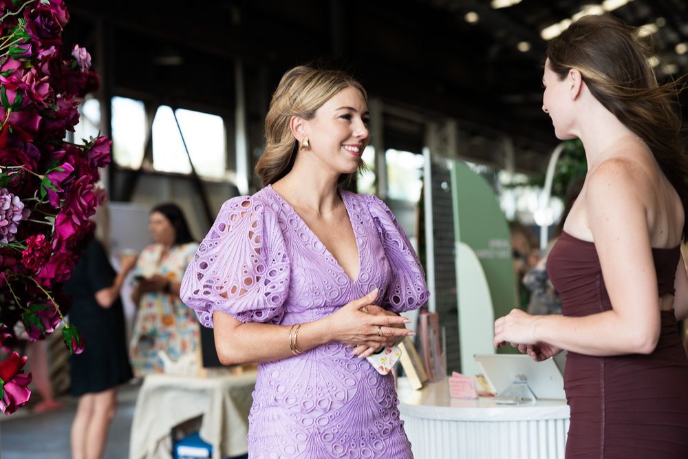 Two women in elegant dresses smiling and conversing at an indoor floral event or wedding reception.