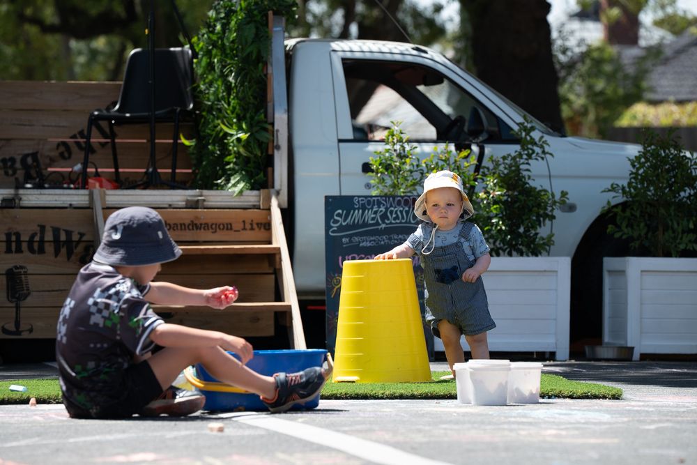 Two toddlers playing outdoors near a white van with a yellow bucket at a summer market.