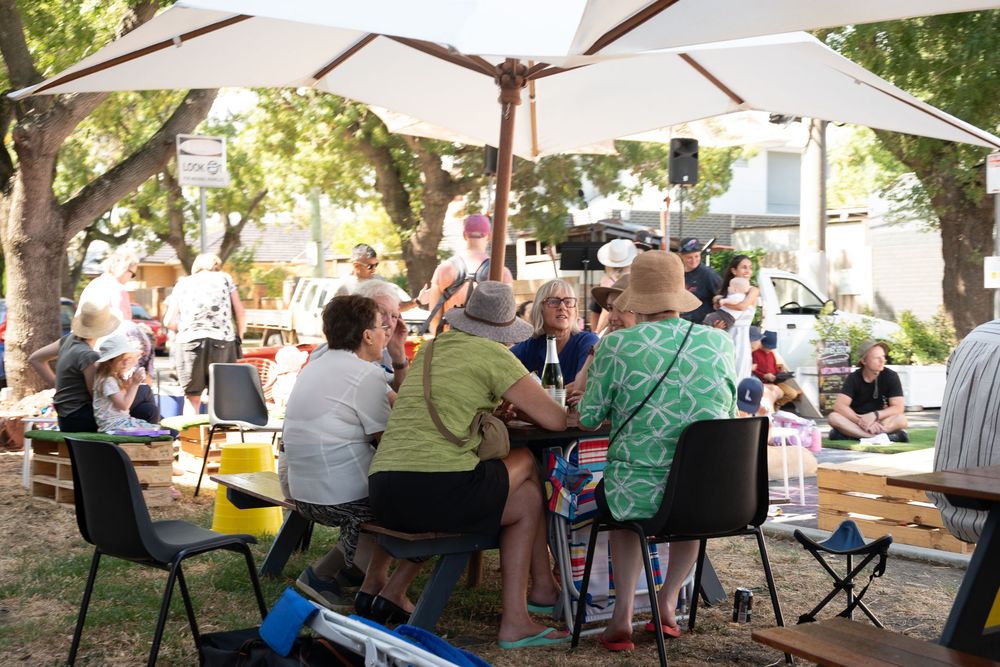 Group of adults sitting together under large umbrellas at an outdoor community gathering on a sunny day.