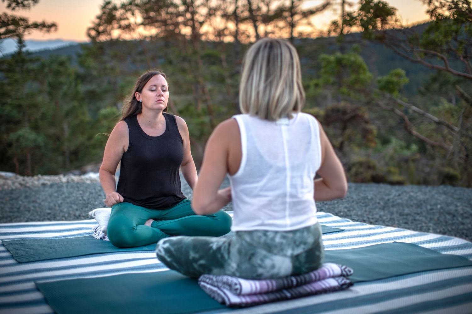 Two women practicing outdoor meditation on yoga mats at sunset surrounded by nature.