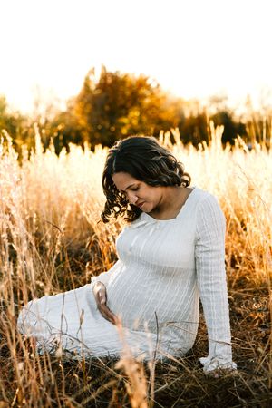 An African-American woman poses in a white dress in a field during her fall maternity photoshoot at Catoosa Sunflower Farm.