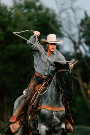 a high school senior cowboy on his blue roan horse run across a pasture swinging a rope for senior photos on family land in Ada, Oklahoma.