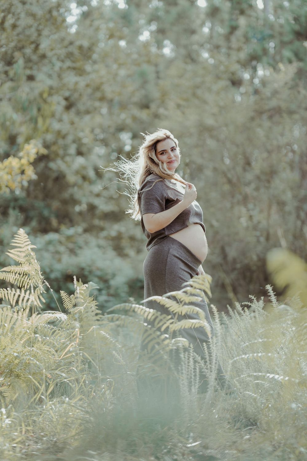 Smiling pregnant woman in gray dress standing among ferns and greenery in a sunlit forest.