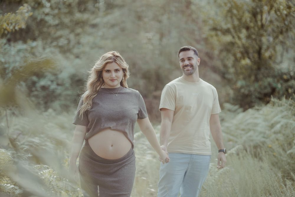 Pregnant couple holding hands walking on a lush green forest path during a maternity photoshoot.