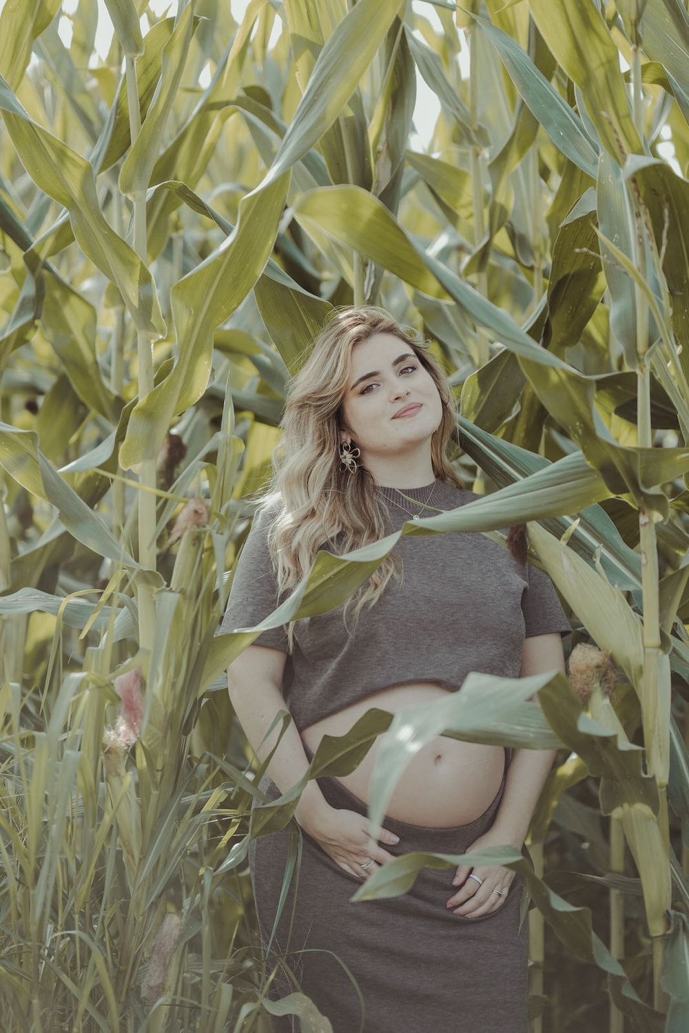 Pregnant woman in gray crop top and skirt standing among tall green corn stalks in a field.