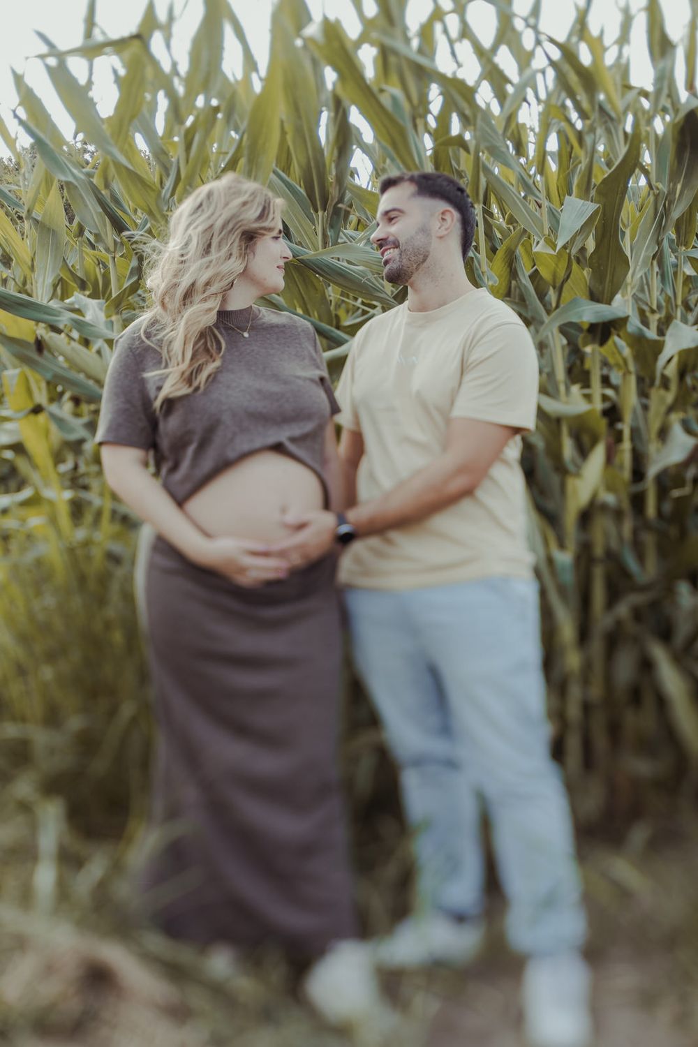 A pregnant couple smiles at each other in front of a lush cornfield, the man gently holding the woman's baby bump.