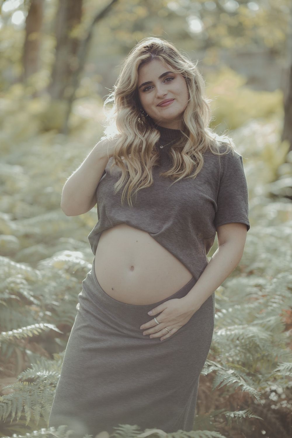 Smiling pregnant woman in gray crop top and skirt cradling baby bump outdoors among ferns in forest.