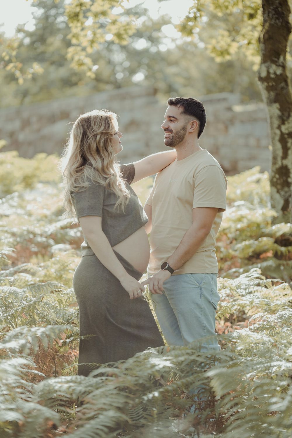 Pregnant couple holding hands in a sunlit fern forest, smiling at each other during a maternity photoshoot.
