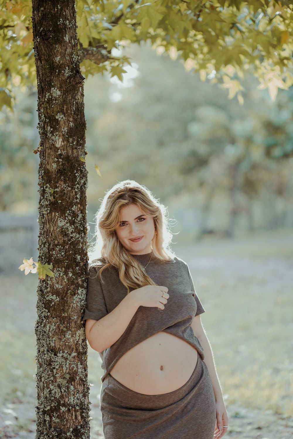 Pregnant woman in gray dress lifting shirt to show baby bump while leaning against tree in sunny park.