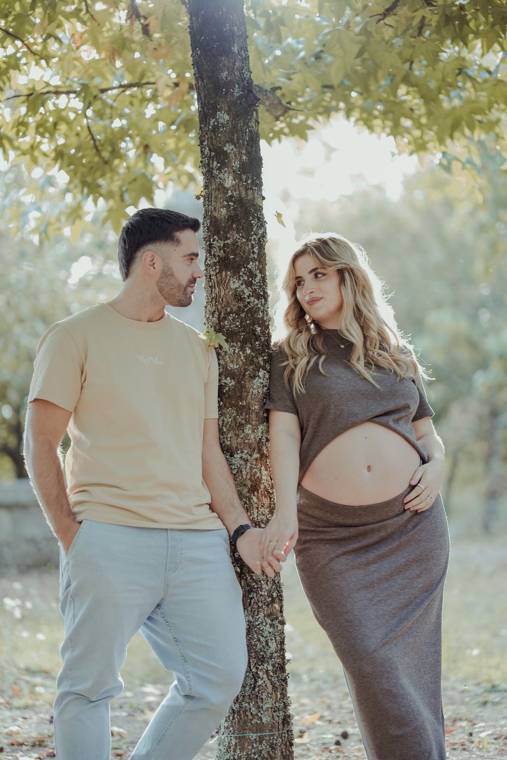 Couple holding hands by a tree in autumn park, pregnant woman in gray dress with partner.