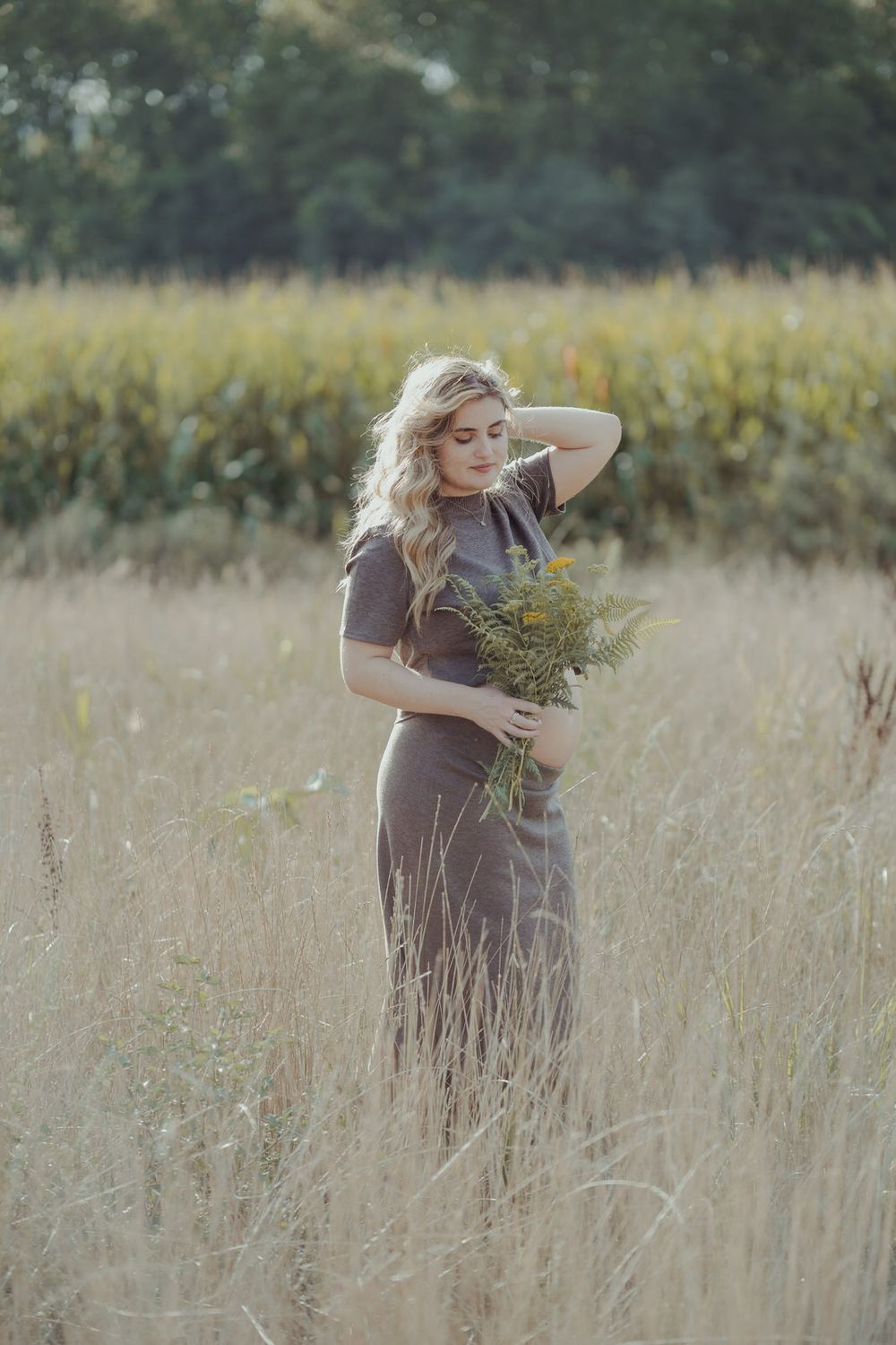 Blonde woman in gray dress holding wildflowers standing in tall grass field with corn in background.
