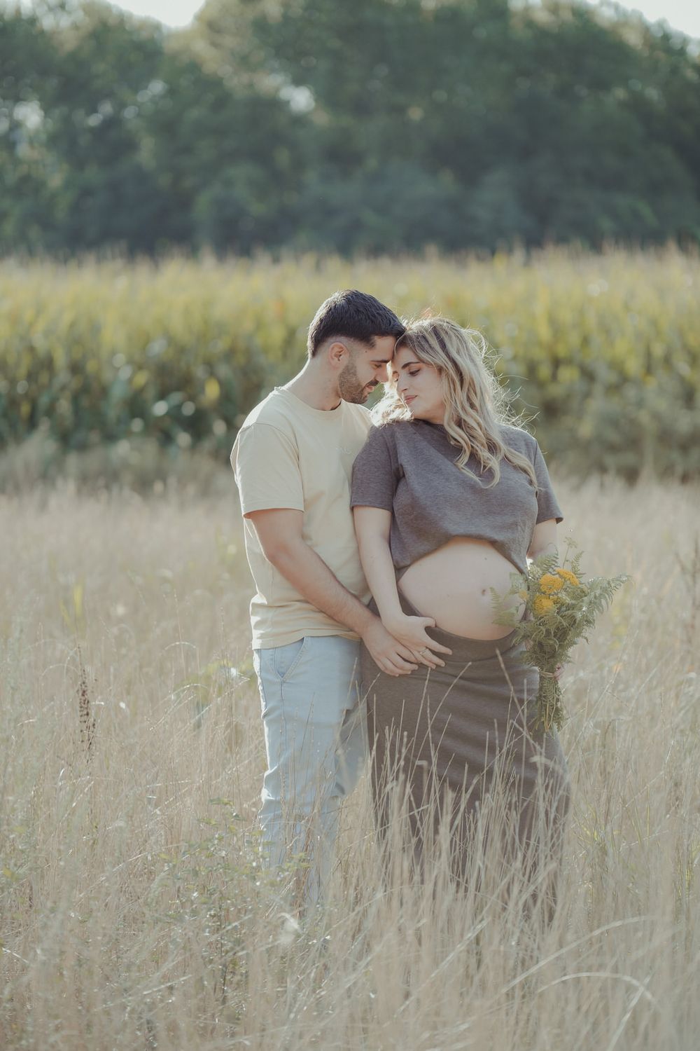 Couple in a golden field, pregnant woman cradling baby bump while partner embraces her tenderly.