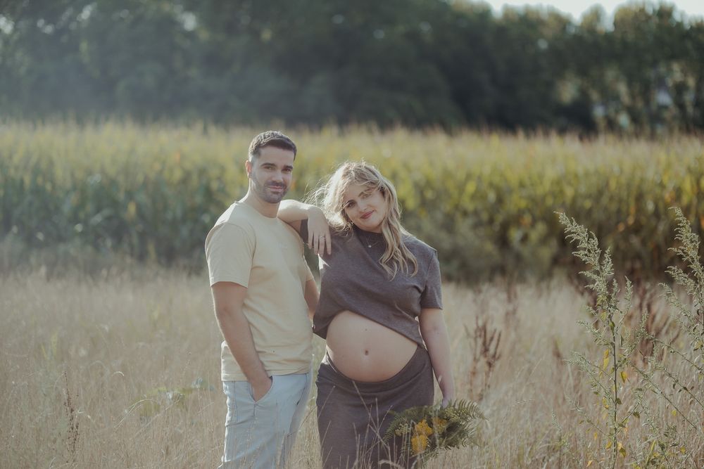 Couple posing in golden field, pregnant woman in gray dress leaning on mans shoulder outdoors.