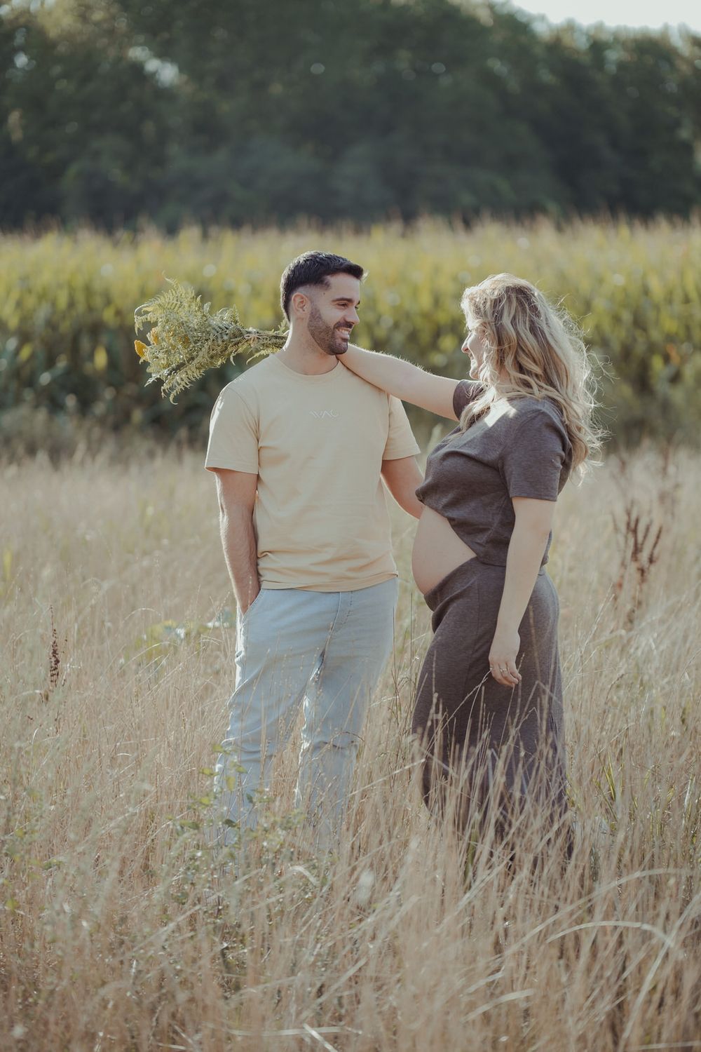 Smiling man in beige shirt with pregnant partner in brown dress standing in sunny wheat field.
