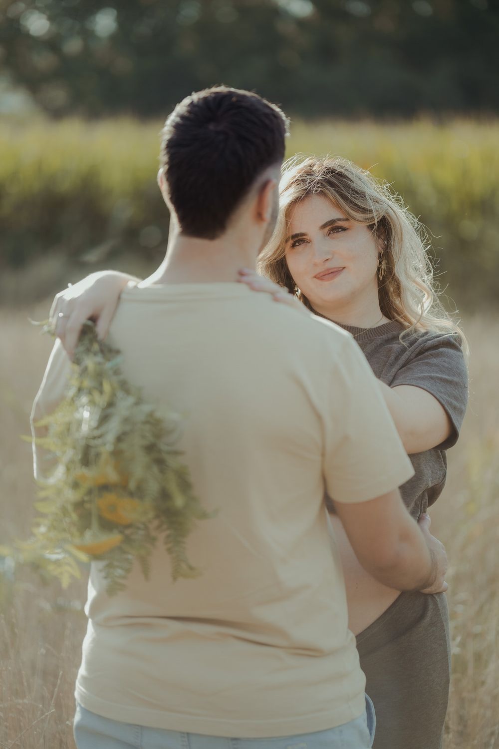 Couple embracing in a golden field, woman holding wildflowers while gazing lovingly at her partner outdoors.