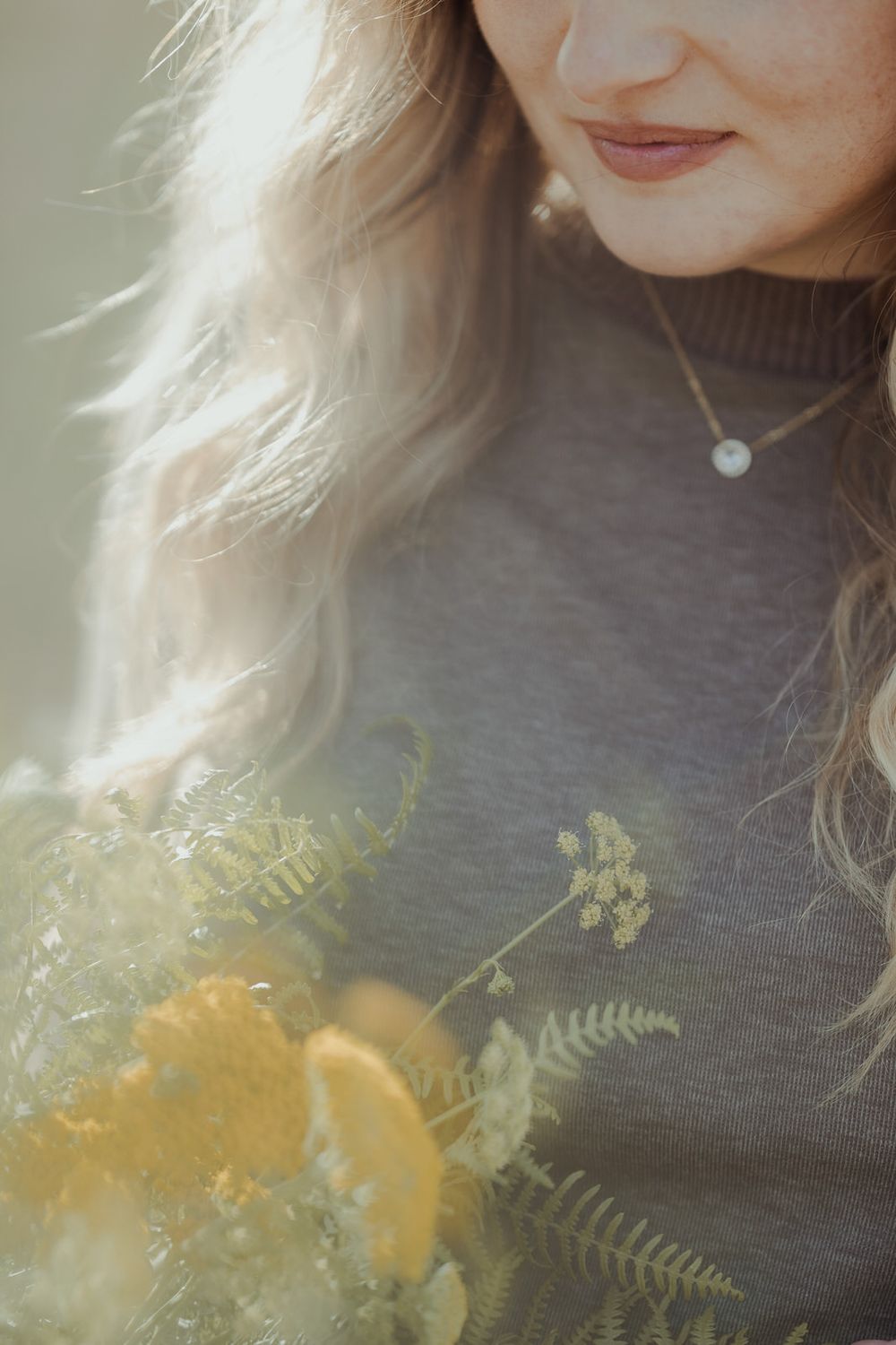 Close-up of blonde woman in gray turtleneck sweater holding yellow flowers wearing pearl necklace.