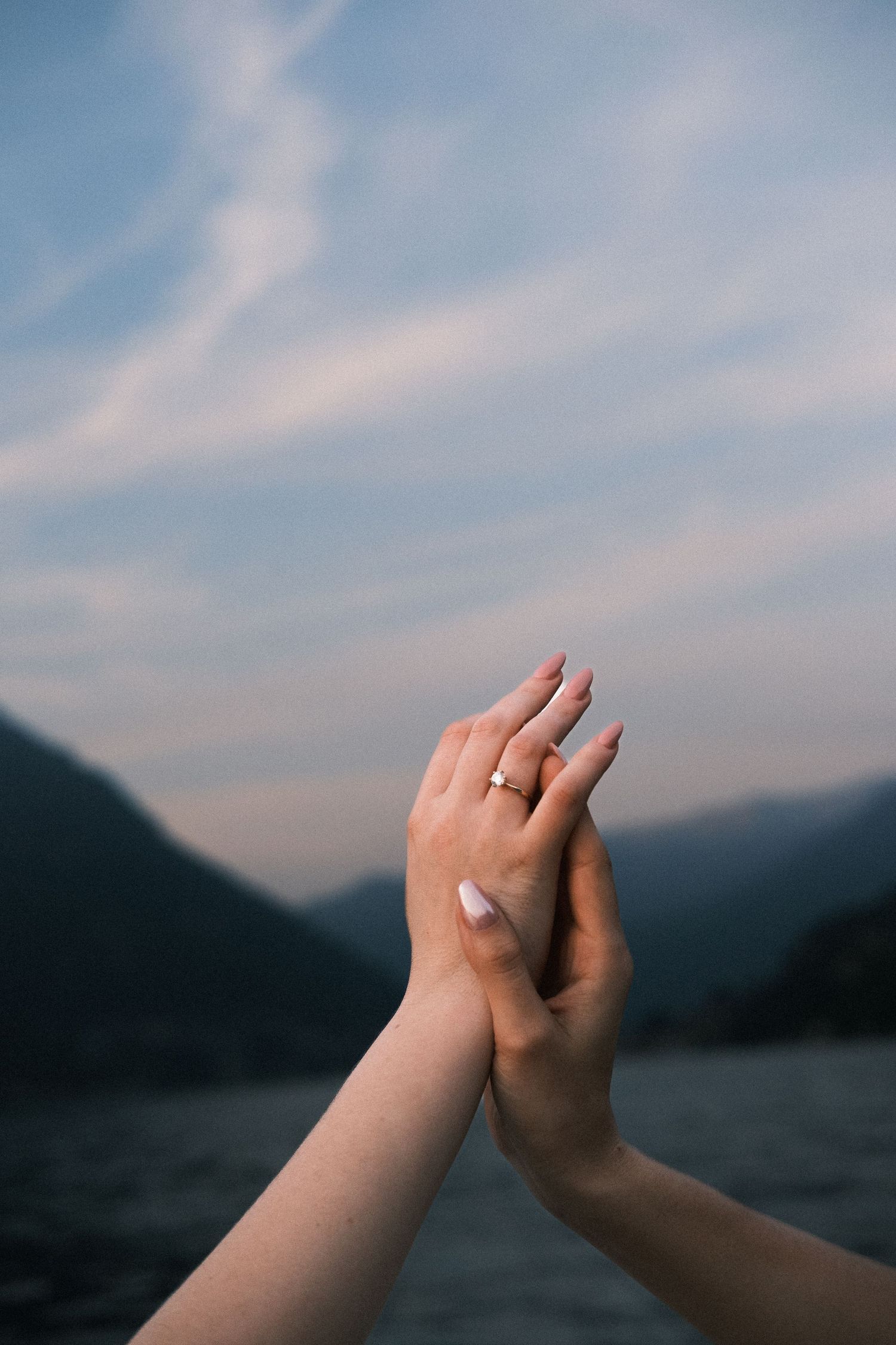 Two hands raised against a mountain lake sky, one wearing a gold engagement ring, symbolizing a romantic proposal.
