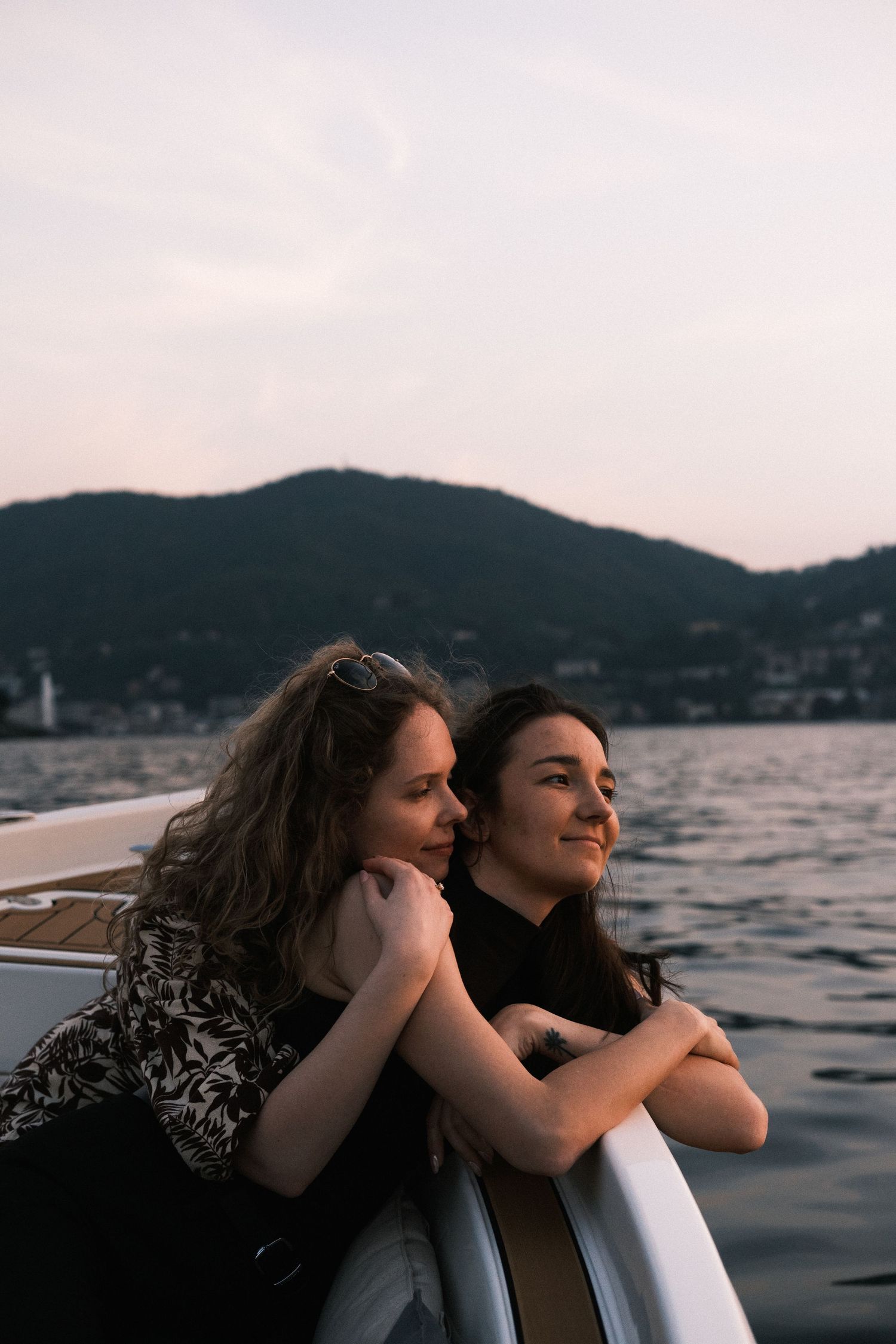 Two women leaning on a boat railing at sunset with mountain lake views in the background.