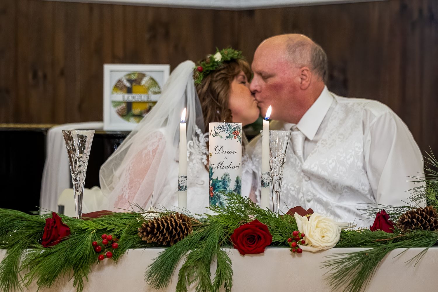 Bride and groom share a kiss at their wedding reception table decorated with pine, red roses, and pinecones.