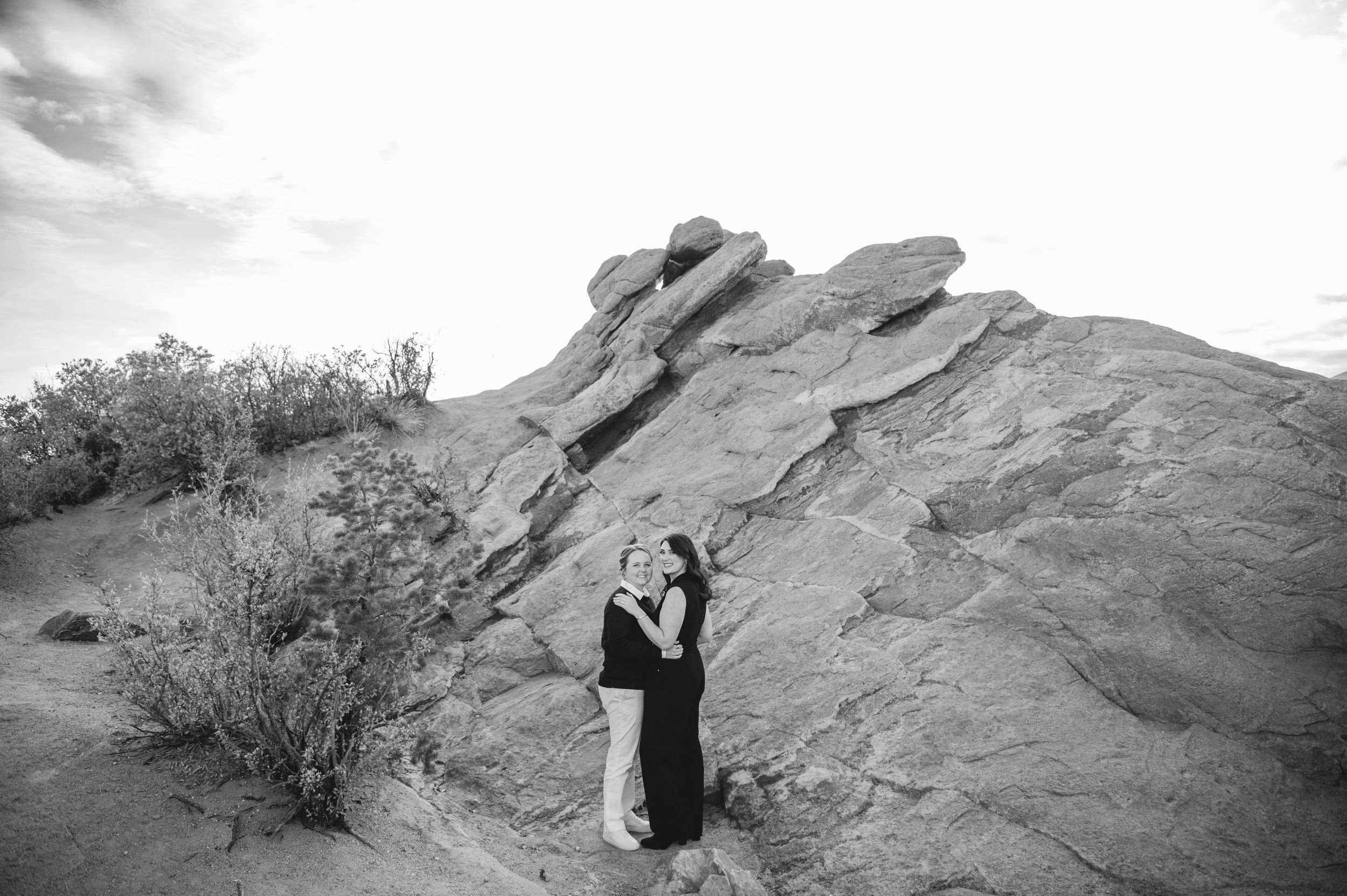 Black and white photo of a couple embracing near large rock formations in a desert landscape.