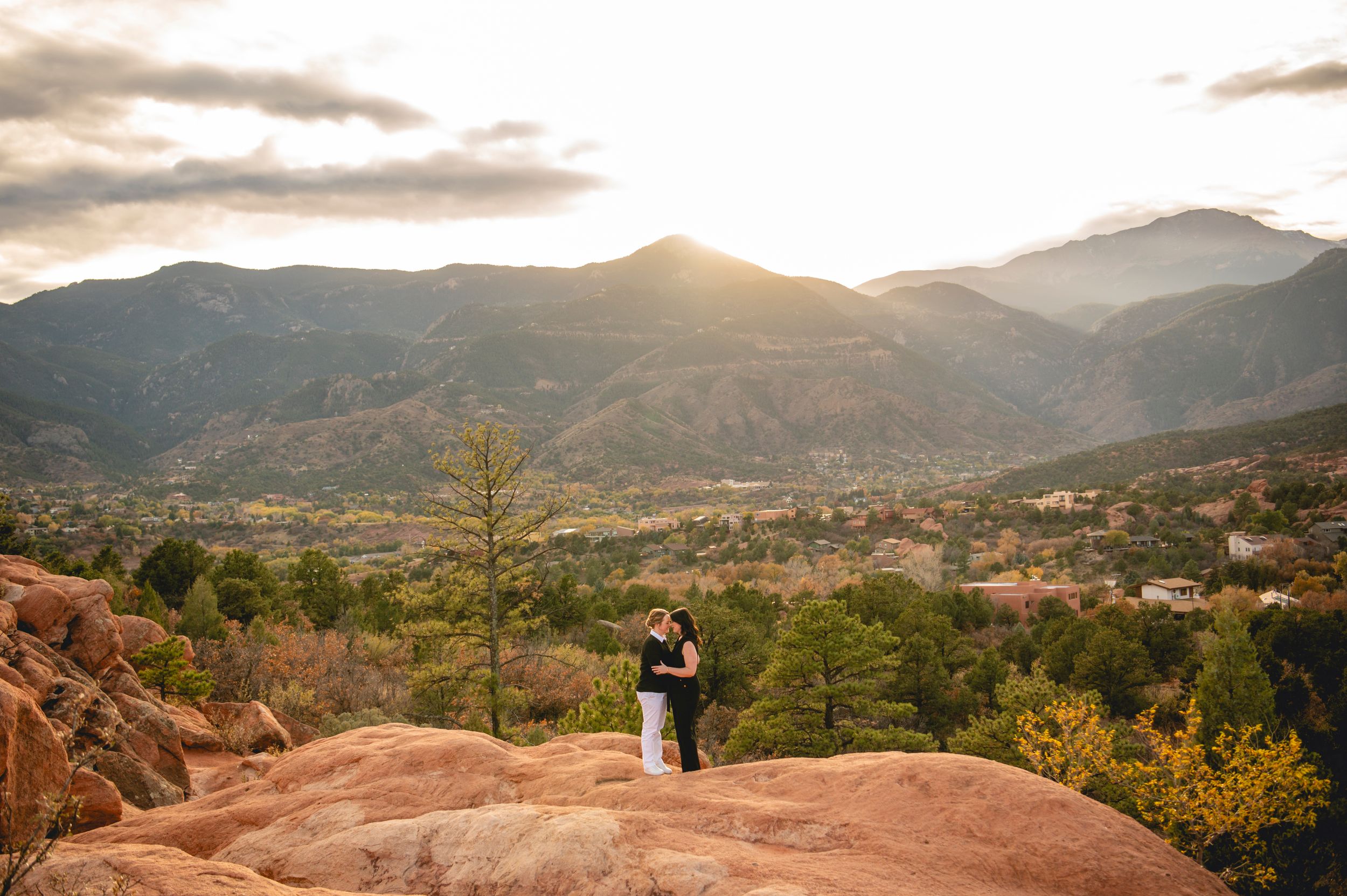 Couple embraces on red rocks at Garden of the Gods with Pikes Peak mountain backdrop at golden sunset.
