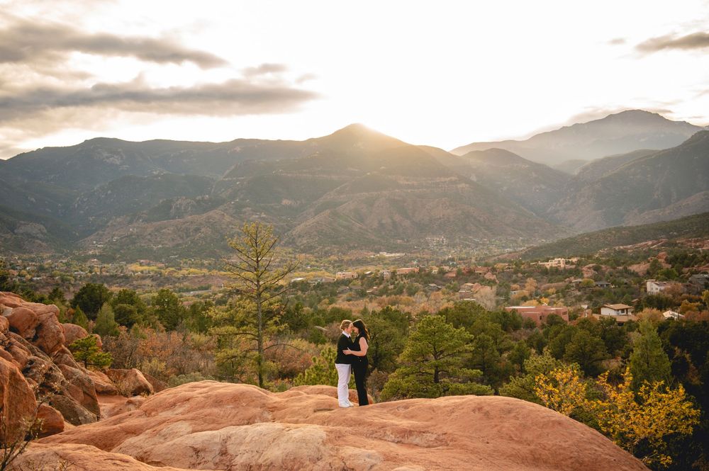 Couple embraces on red rocks at Garden of the Gods with Pikes Peak mountain backdrop at golden sunset.