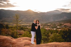 Two women embrace on red rocks at Garden of the Gods with mountain sunset backdrop in Colorado Springs.