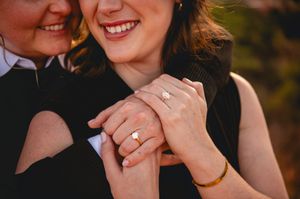 Two women embrace, one displaying an engagement ring and gold bracelet, smiling joyfully in a romantic outdoor setting.