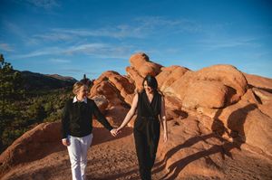 Couple holding hands on red rock formations at Garden of the Gods, Colorado, under a bright blue sky.