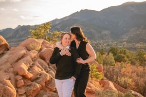 Two women embrace and laugh together at Garden of the Gods with red rocks and mountains at golden hour.