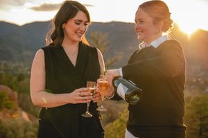 Two women toasting with champagne glasses at sunset with mountain views in the background.