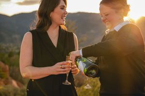 Two women celebrating outdoors at sunset, one pouring champagne into a glass, with mountains in the background.
